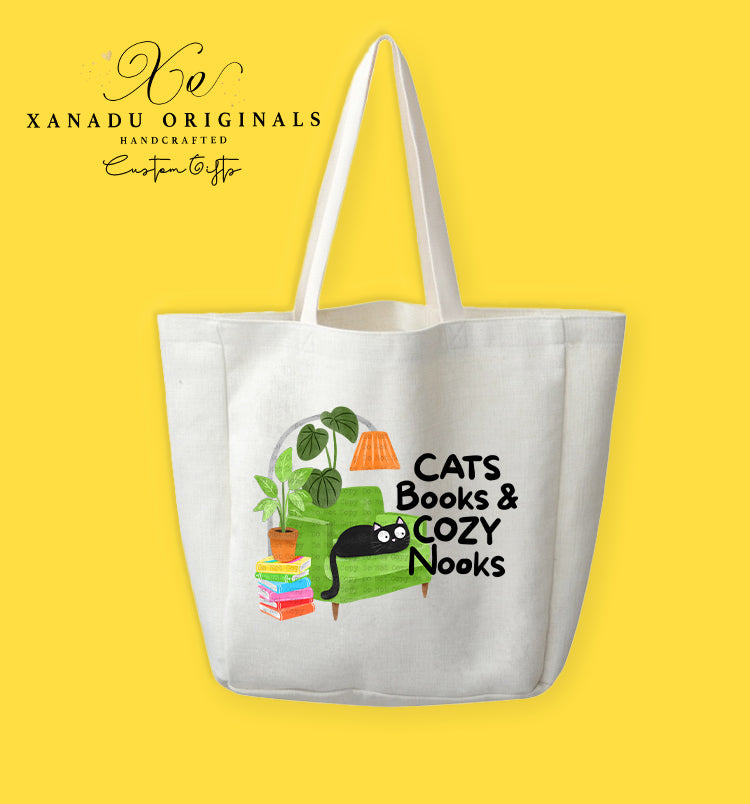 A cream-colored polyester linen tote bag featuring a watercolor illustration of a green armchair, a stack of colorful books, potted plants, and an orange lamp, with a wide-eyed black cat looking out next to text that reads "CATS Books & COZY Nooks."
