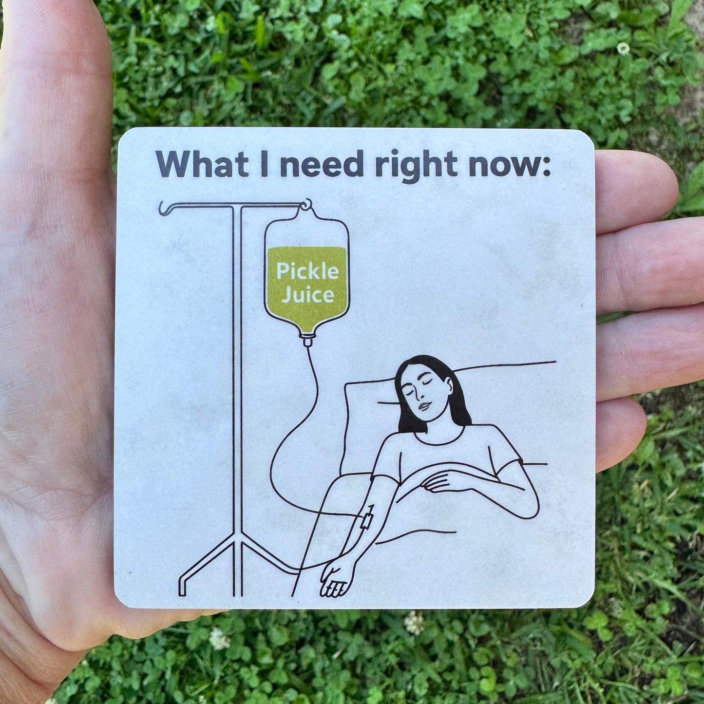 3.6 inch square MDF wood coaster with rounded corners featuring a black line art illustration of a person lying in bed hooked up to an IV pole holding a green bag labeled Pickle Juice, text above reads What I need right now.