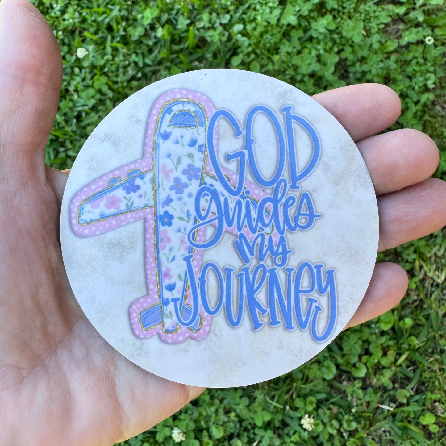 3.6 inch round circular MDF wood coaster featuring a stylized passenger airplane illustration patterned with blue flowers and pink polka dots, next to periwinkle blue typography reading God guides my journey on a rustic cream background.