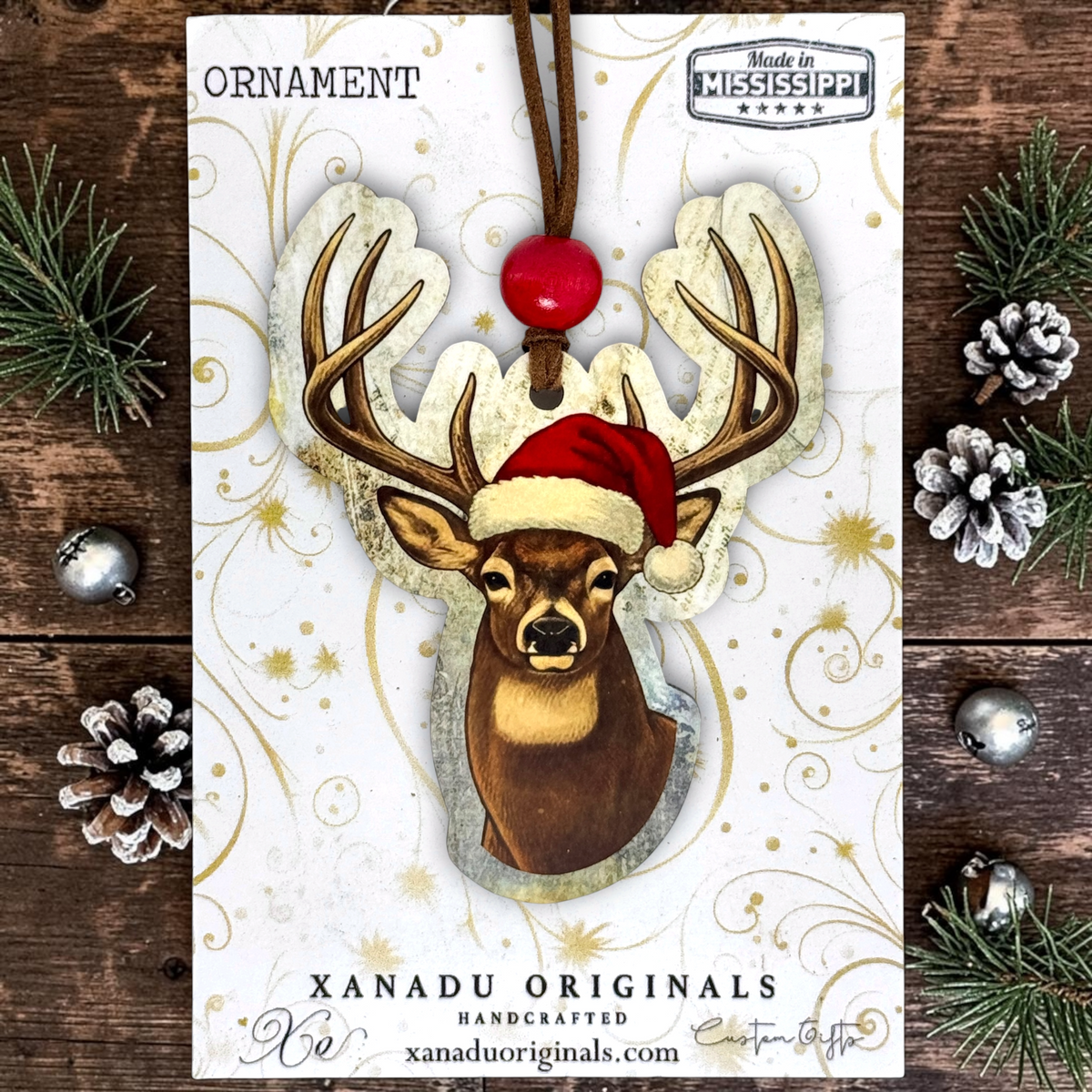 A custom-cut wood ornament showing a deer head in a red Santa hat with a vintage text background, mounted on a gold swirl display card next to pine sprigs and cones.