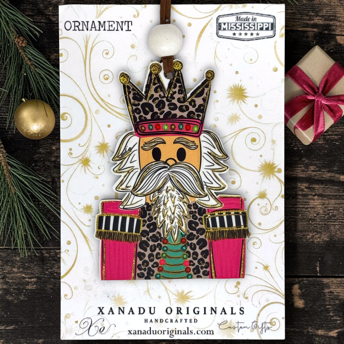 A handcrafted Christmas ornament on its branded display card, styled on a dark wood surface with pine branches and a wrapped gift. The ornament depicts a nutcracker figure with a leopard-print crown, white beard, and pink and green clothing.