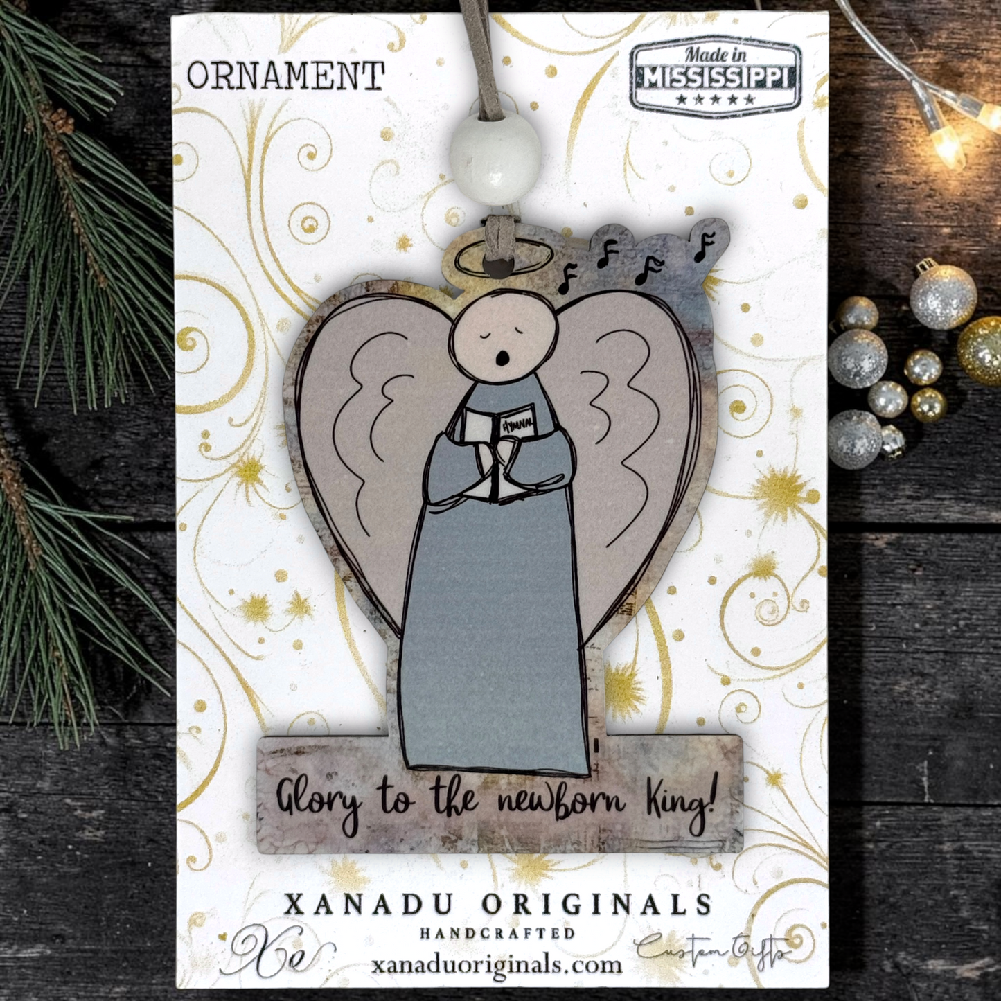 A handmade Christmas ornament by Xanadu Originals, featuring a stylized angel figure with a gray-blue robe and wings, set against a heart-shaped background. The ornament is mounted on a decorative display card with gold swirls and text reading 'ORNAMENT' and 'glory to the newborn king!'.