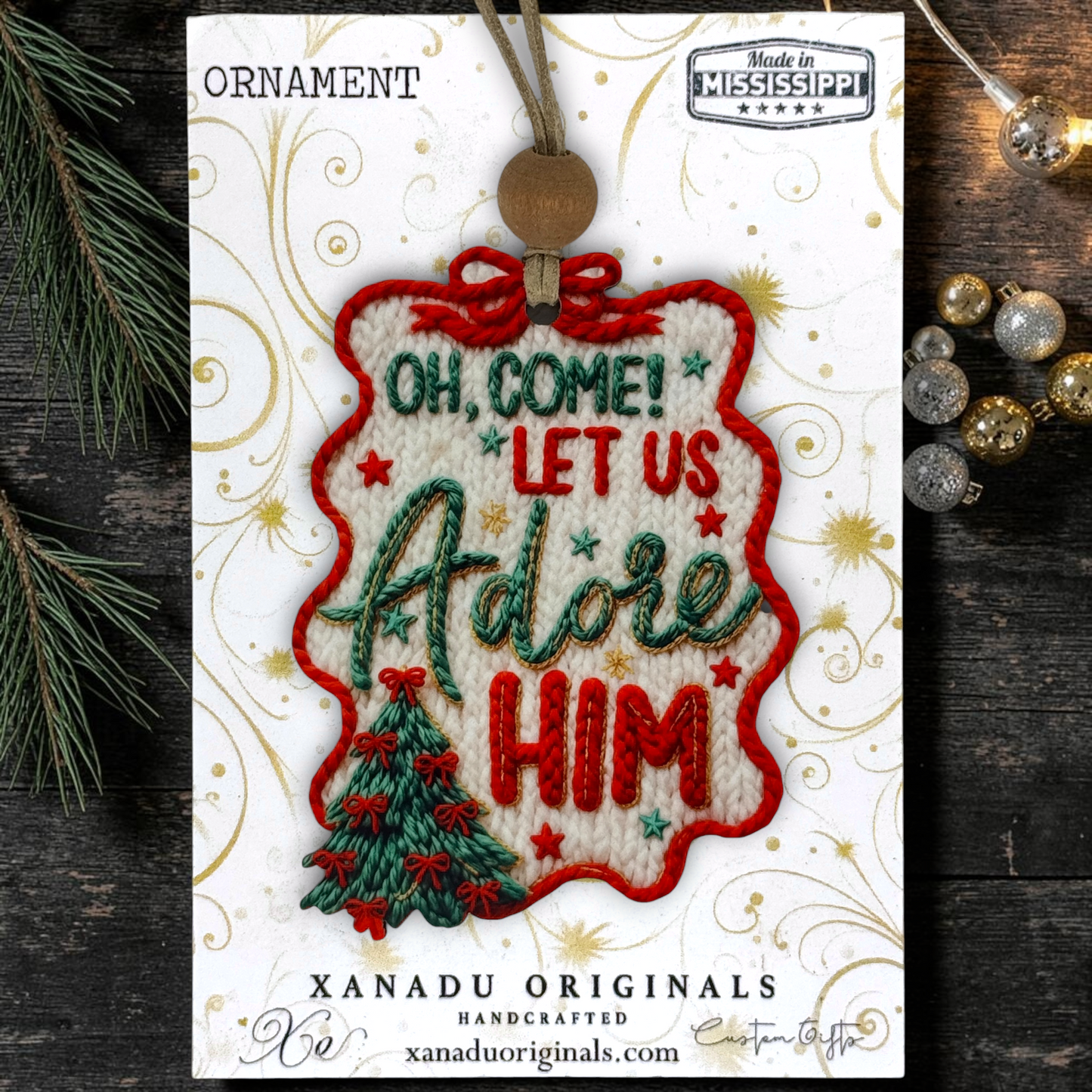 A Christmas ornament package from Xanadu Originals featuring a white embroidered ornament with red and green lettering that reads 'Oh, Come! Let Us Adore Him' and a small green tree at the bottom. The package is displayed on a dark wood surface with pine branches and silver/gold ornaments around it.