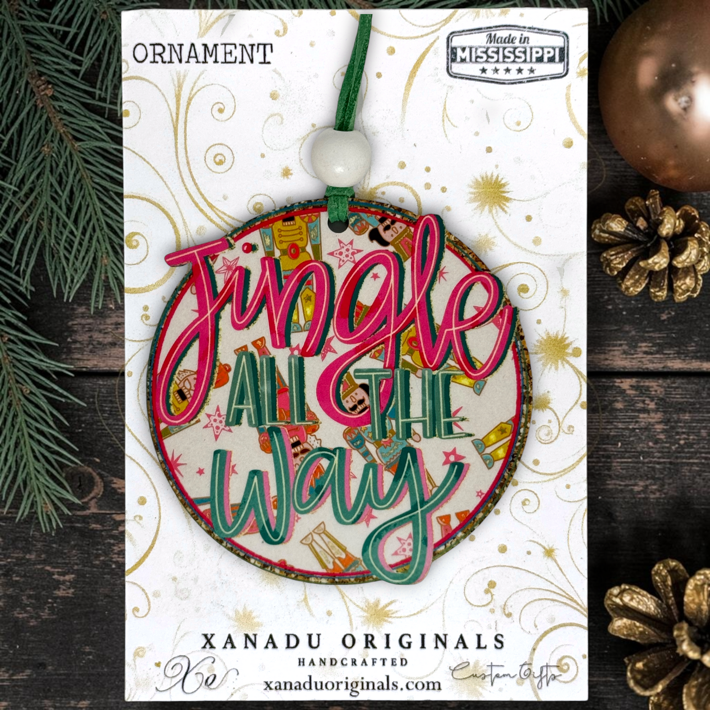 A round Christmas ornament carded on a branded display card. The ornament has layered script lettering reading 'Jingle All The Way' in pink and green, with a festive collage background and a green hanging cord.