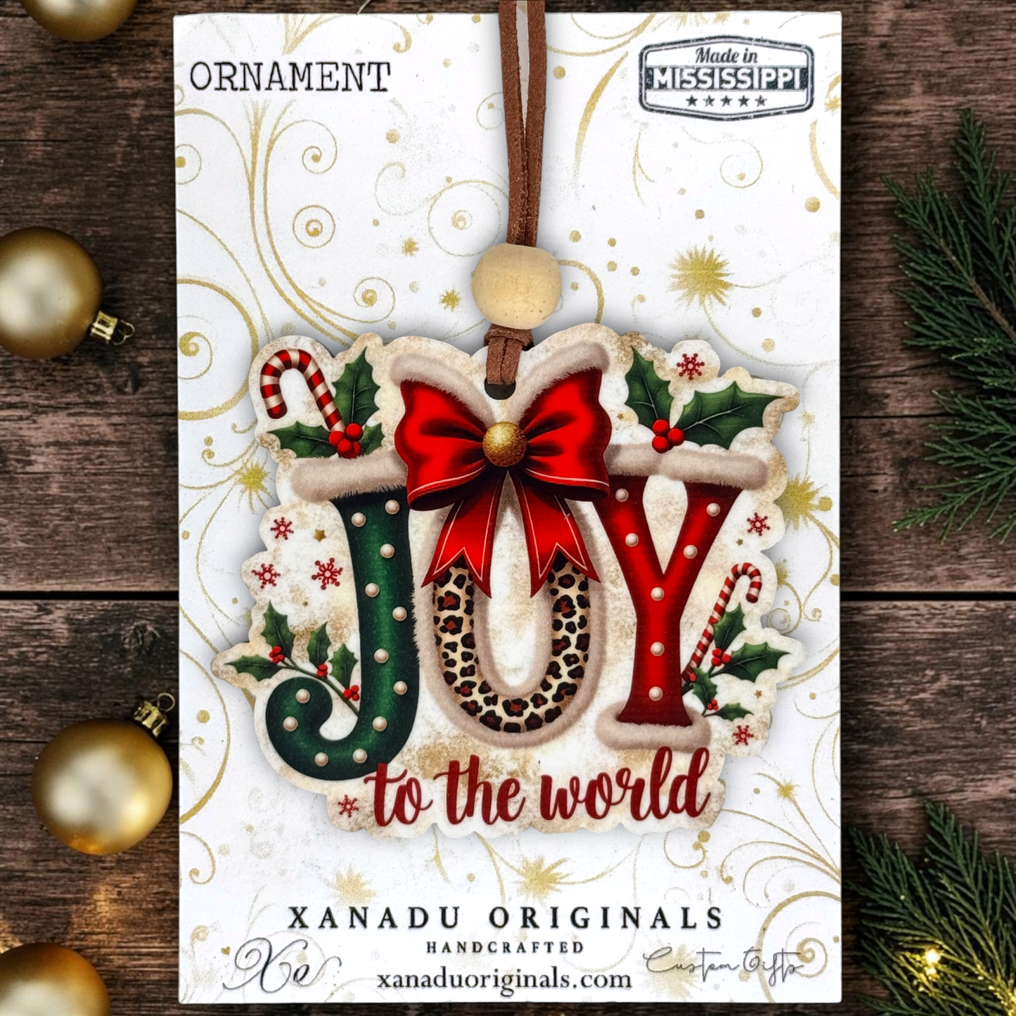 A festive Christmas ornament on a card featuring a handcrafted ornament that spells 'JOY to the world' with a red bow, leopard print letter 'O', and holiday accents. The card is displayed on a rustic wooden background with gold ornaments and evergreen branches.