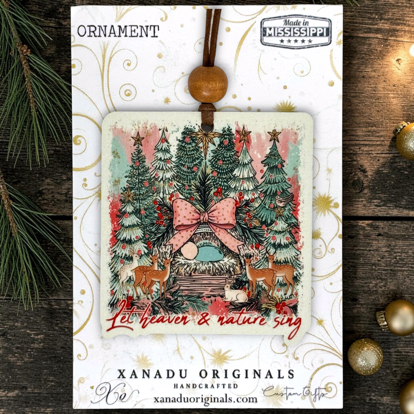 A square Christmas ornament card with a hanging wooden bead and ribbon, featuring a festive illustrated nativity scene with pine trees, a pink bow, deer, and the text 'Let heaven & nature sing.' The card is branded XANADU ORIGINALS and photographed on a dark wood surface with holiday decorations around it.