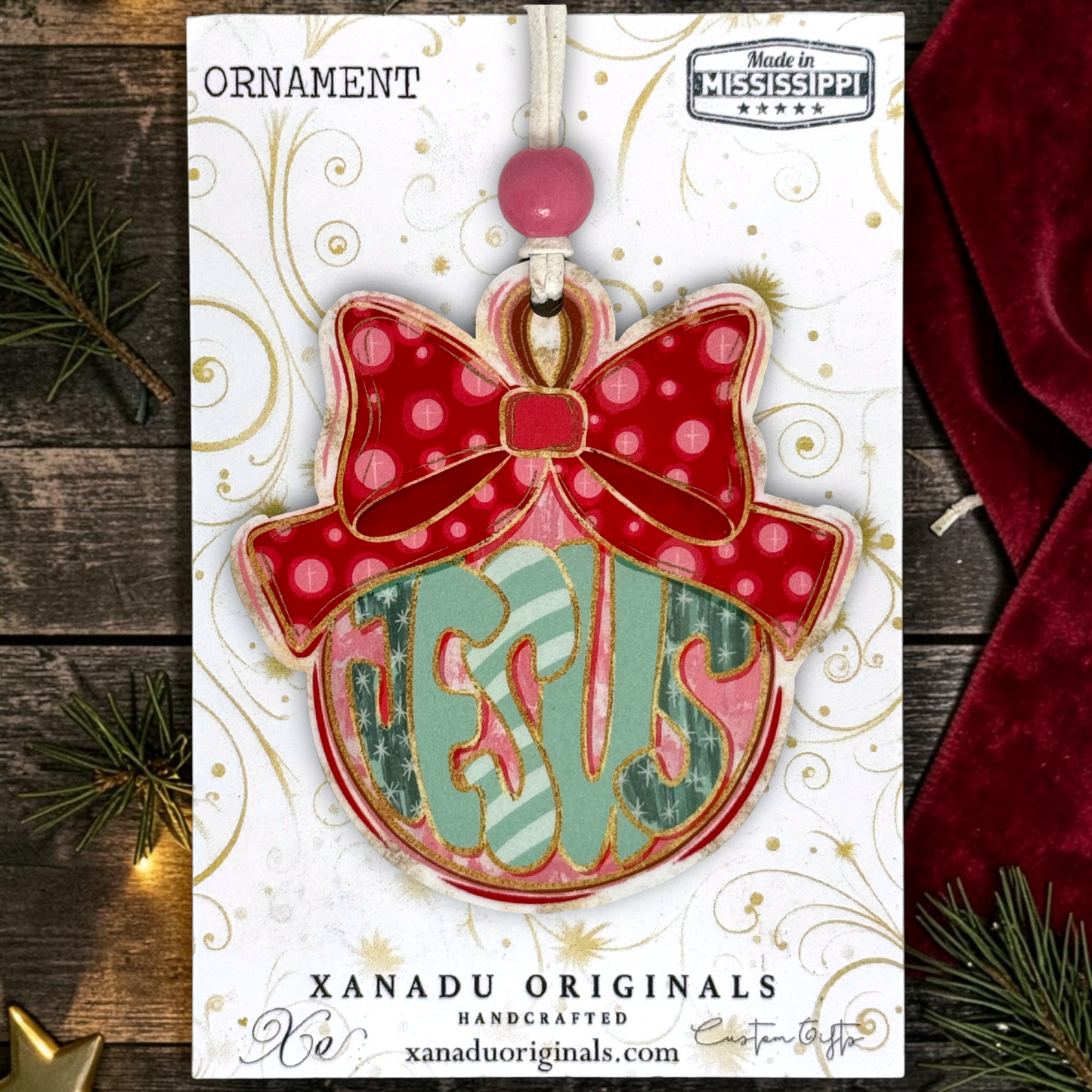 A Christmas ornament in its retail packaging, featuring a red bow with pink polka dots and a round ornament body with the word 'JESUS' in green and pink striped letters. The packaging card is displayed on a rustic wooden background with pine branches and a red fabric corner.