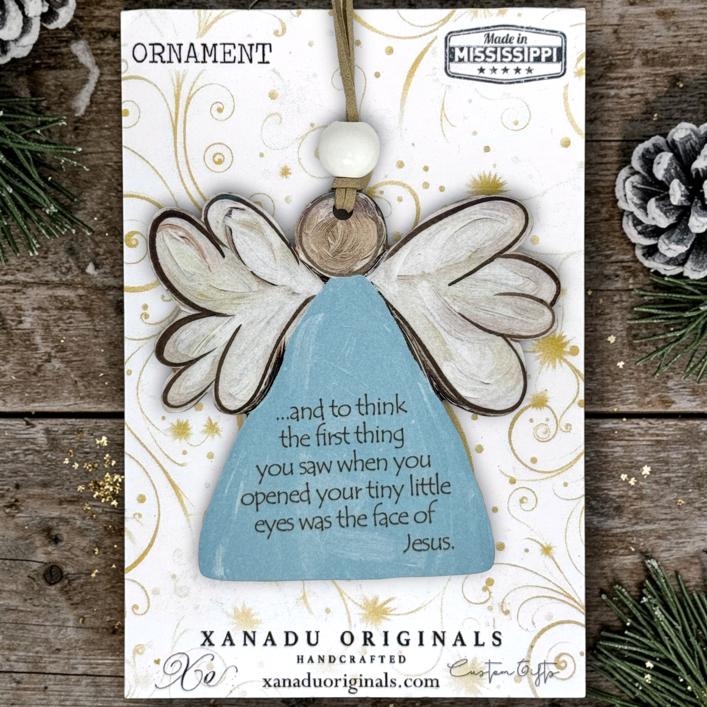 Handcrafted born sleeping angel ornament by Xanadu Originals, painted in light blue and white with brown outlines, displayed on its white printed backing card over a rustic wood surface with pine branches and a pinecone.