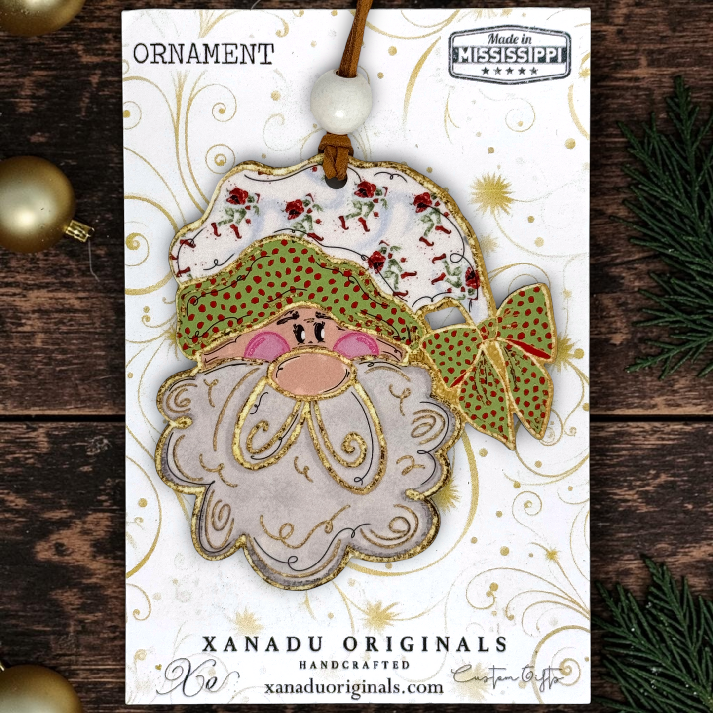 A handmade Christmas ornament card featuring a Santa Claus face ornament with a patterned hat, green polka-dot bow, and white beard, displayed on a branded card over a rustic wood background with holiday decorations.