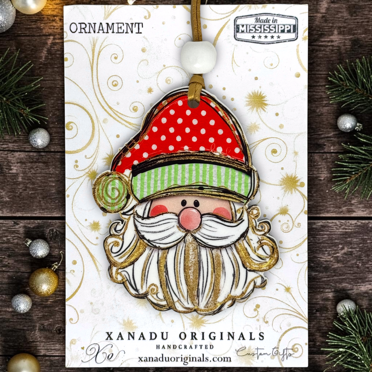 A festive Christmas ornament featuring a handcrafted Santa Claus face with a red polka-dot hat, green striped trim, white beard, and gold glitter details, hanging from a brown ribbon on a white card. The card is photographed on a dark wood surface with holiday greenery and ornaments around it.