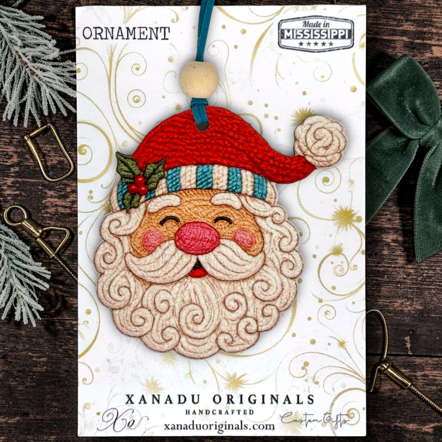 A festive wooden faux embroidered Santa Claus ornament on a card from Xanadu Originals, featuring a 3D stitched Santa face with a red hat, blue striped trim, and cream beard, displayed on a dark wood background with holiday props.