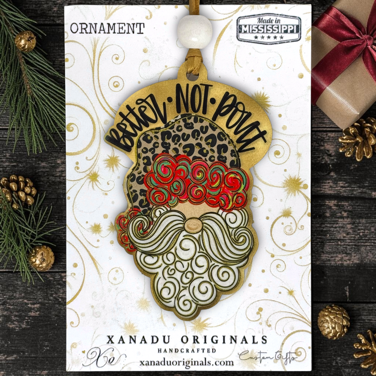 A leopard print "You Better Not Pout" Santa wood ornament displayed flat on a white Xanadu Originals packaging card featuring elegant gold swirl patterns.