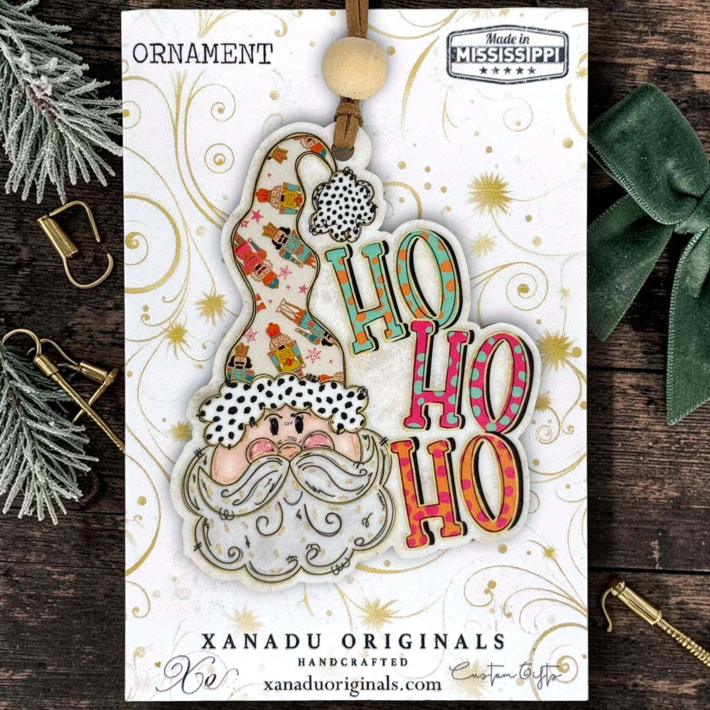 A festive Christmas ornament card from Xanadu Originals, featuring a Santa Claus ornament with a patterned hat and colorful 'HO HO HO' lettering, displayed on a dark wood background with holiday props.