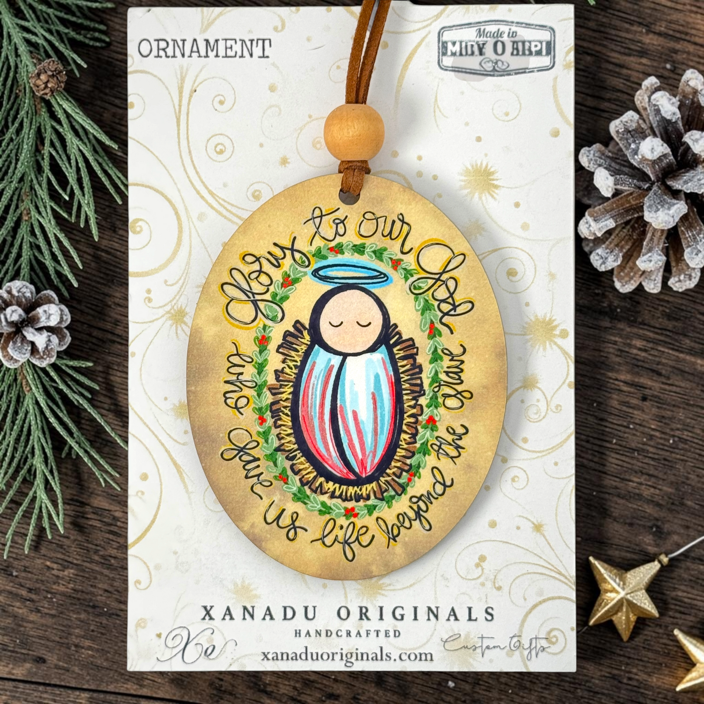 Handcrafted oval Christmas ornament featuring a baby Jesus illustration with a halo and the text 'Love to our God Your Life gives us life because the Christ', displayed on its branded card against a rustic pine and wood background.