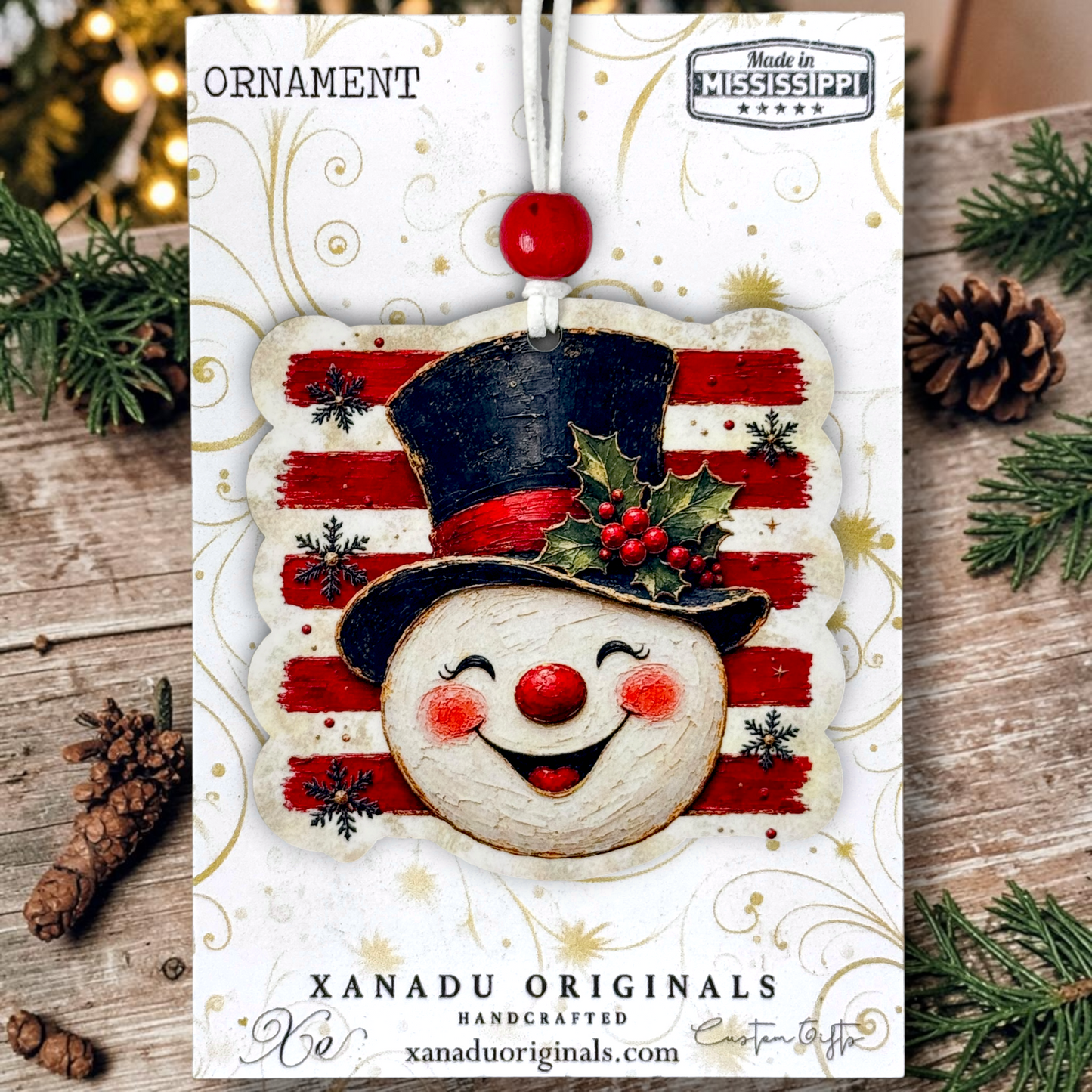 A festive Christmas ornament card featuring a handcrafted snowman ornament with a black top hat, red nose, red cheeks, and a holly accent on a red-and-white striped background. The card is displayed on a rustic wooden surface with pine branches, pinecones, and warm blurred lights around it.