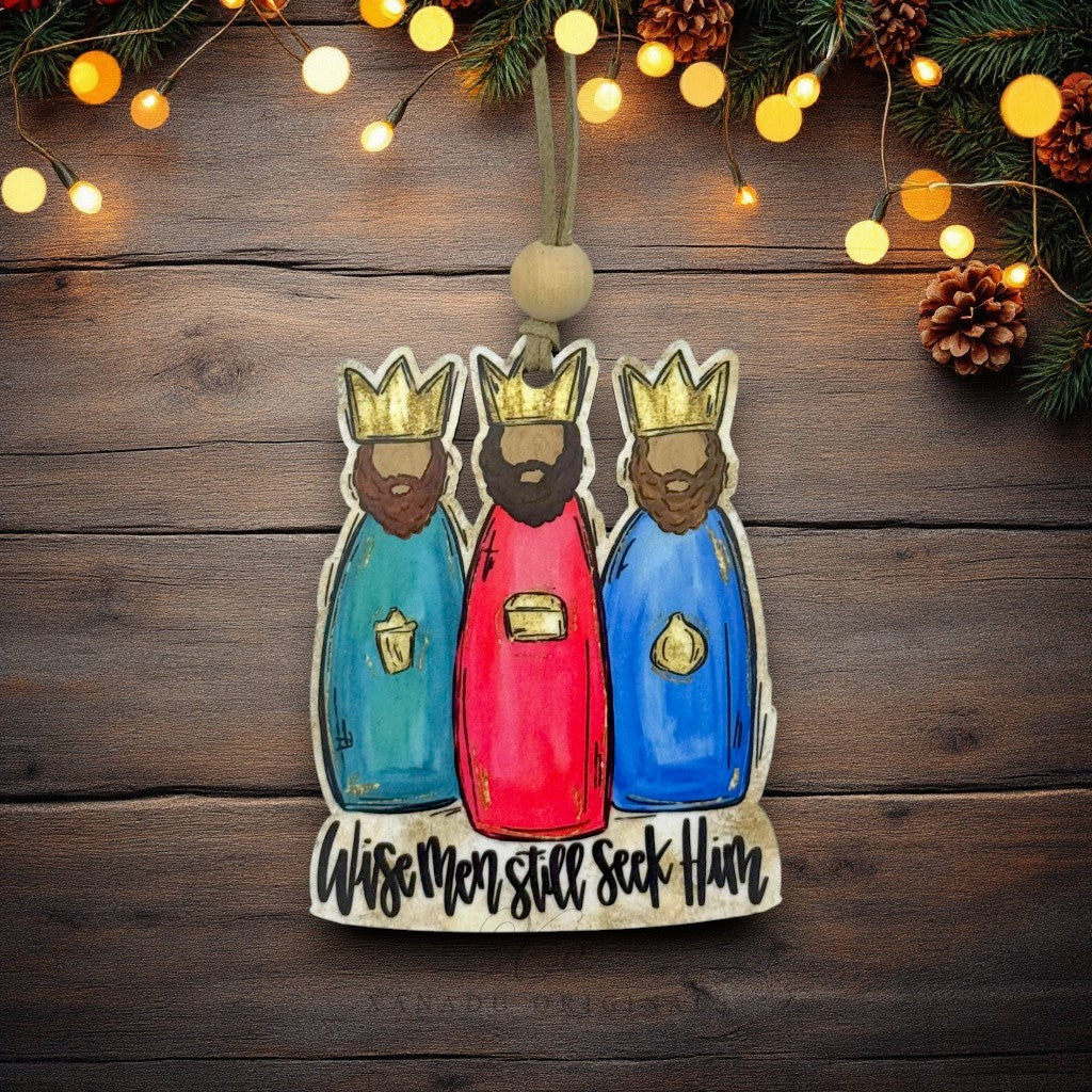 A hand-painted wooden Christmas ornament depicting the Three Wise Men, hanging against a rustic dark wood background decorated with pine branches, pinecones, and warm string lights.