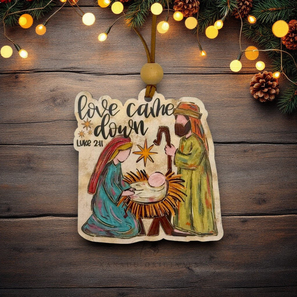 A hand-painted wooden Christmas ornament featuring the Nativity scene with Mary, Joseph, baby Jesus in a manger, and a star, with the text 'love came down Luke 2:11'. The ornament is hanging from a wooden string with a round bead, set against a rustic dark wood background decorated with pine branches, pinecones, and warm glowing fairy lights.