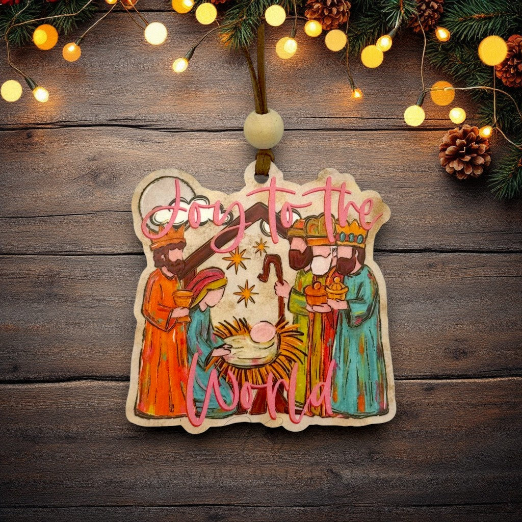 A hand-painted wooden Christmas ornament featuring Nativity figures and the words 'Joy to the World' in pink script, hanging against a rustic dark wood background with warm string lights and pine branches.