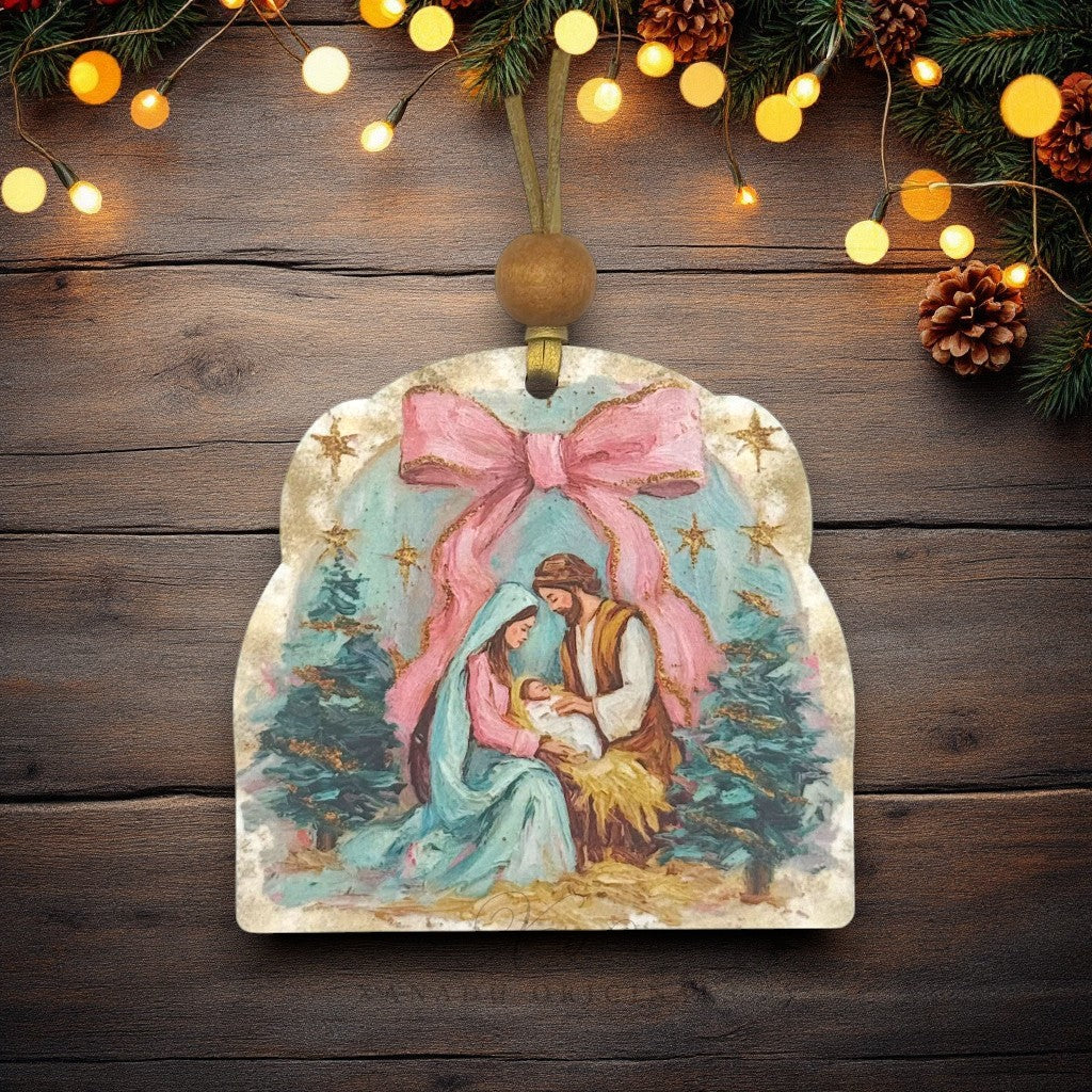 A Christmas ornament shaped like a rustic wooden sign with a painted nativity scene featuring Mary, Joseph, and baby Jesus under a large pink bow, hanging on a dark wood background with warm string lights and pine branches.