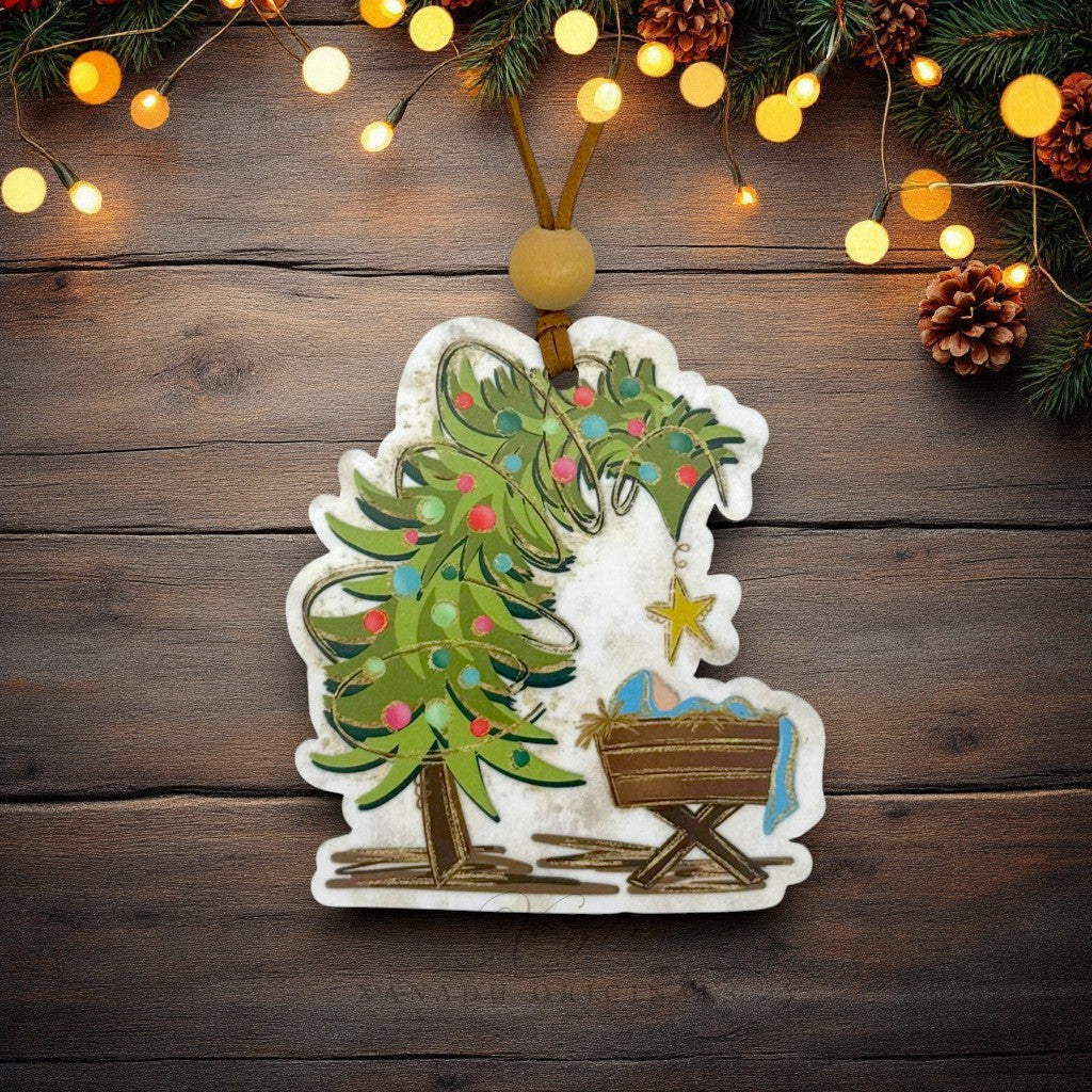 A handcrafted Christmas ornament featuring a green decorated Christmas tree and a wooden-style baby cradle with a blue blanket and gold star, hanging on a rustic dark wood background with warm string lights and pine branches.