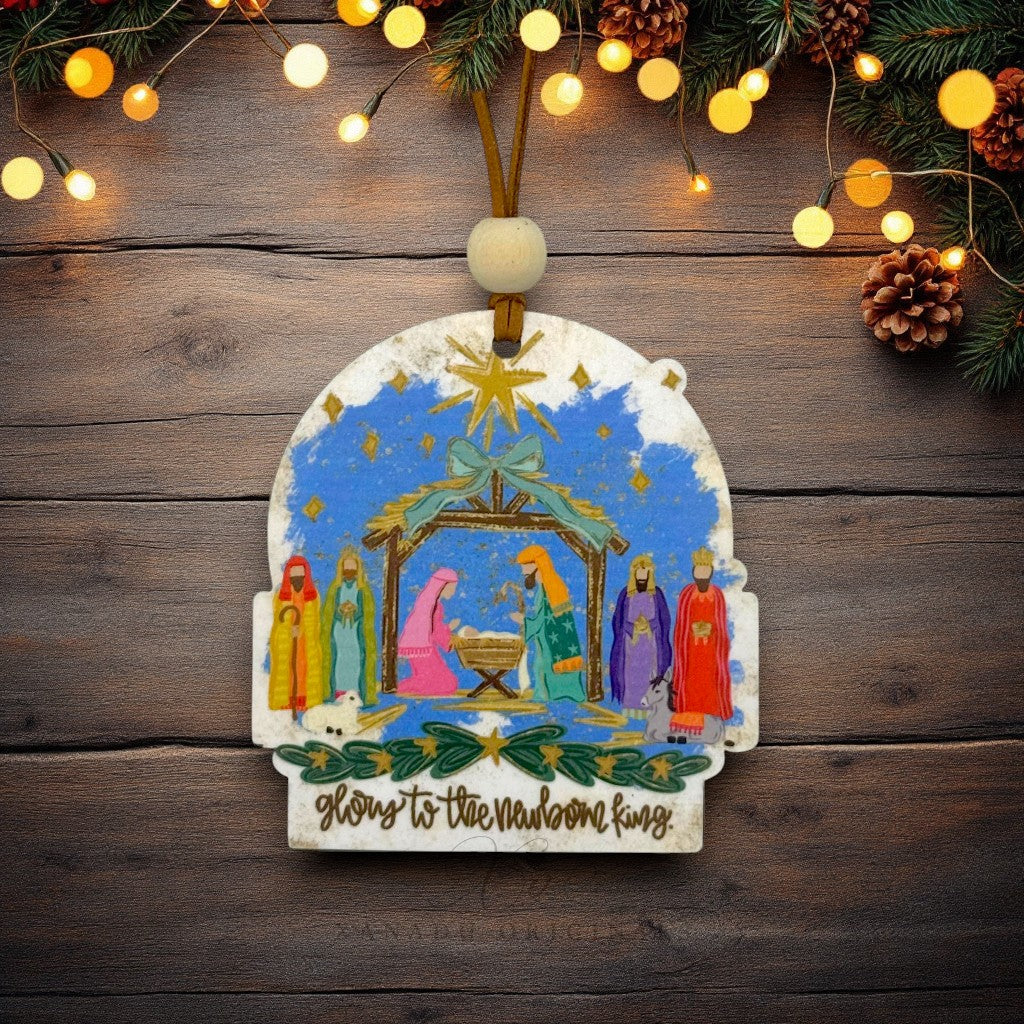 A hand-painted wooden Christmas nativity ornament hanging on a rustic dark wood background with warm string lights and pine garlands. The ornament features a colorful nativity scene with Mary, Joseph, baby Jesus, and wise men under a star, with 'Glory to the newborn king' written at the bottom.