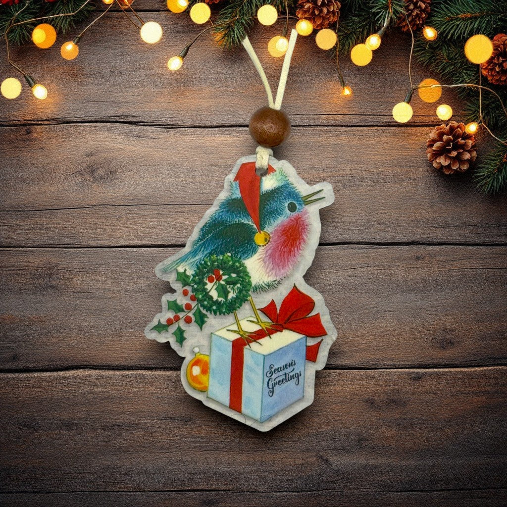 A Christmas bird-shaped ornament with a robin design, a red Santa hat, green wreath, holly, gift box, and 'Season's Greetings' text, hanging on a rustic wooden background with warm string lights and pine branches.