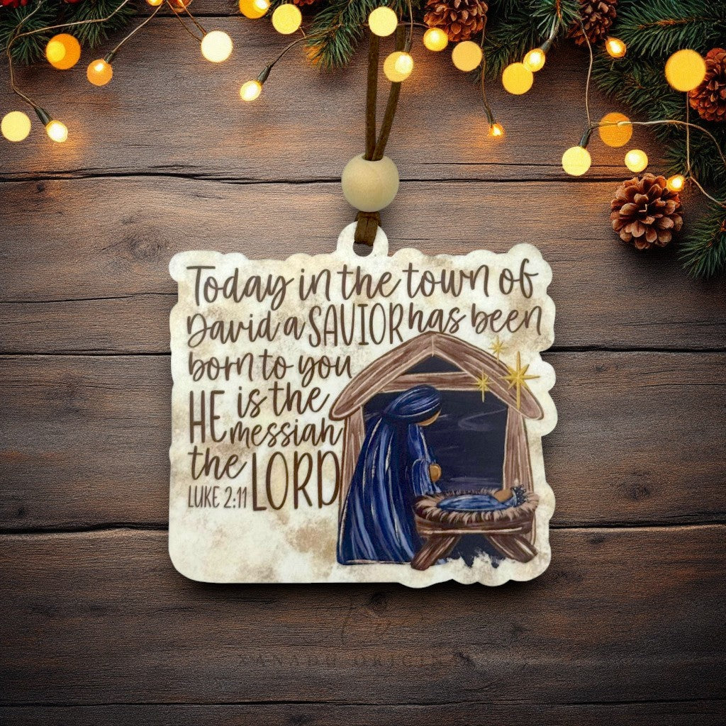 A Christmas ornament featuring a hand-lettered Bible verse and an illustration of the Holy Family in a stable, hanging from a wooden surface decorated with pine branches and warm string lights.