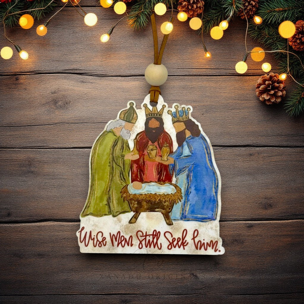 A hand-painted wooden Christmas ornament depicting the Three Wise Men and Baby Jesus in a manger, hanging on a rustic dark wood background with warm string lights and pine branches.