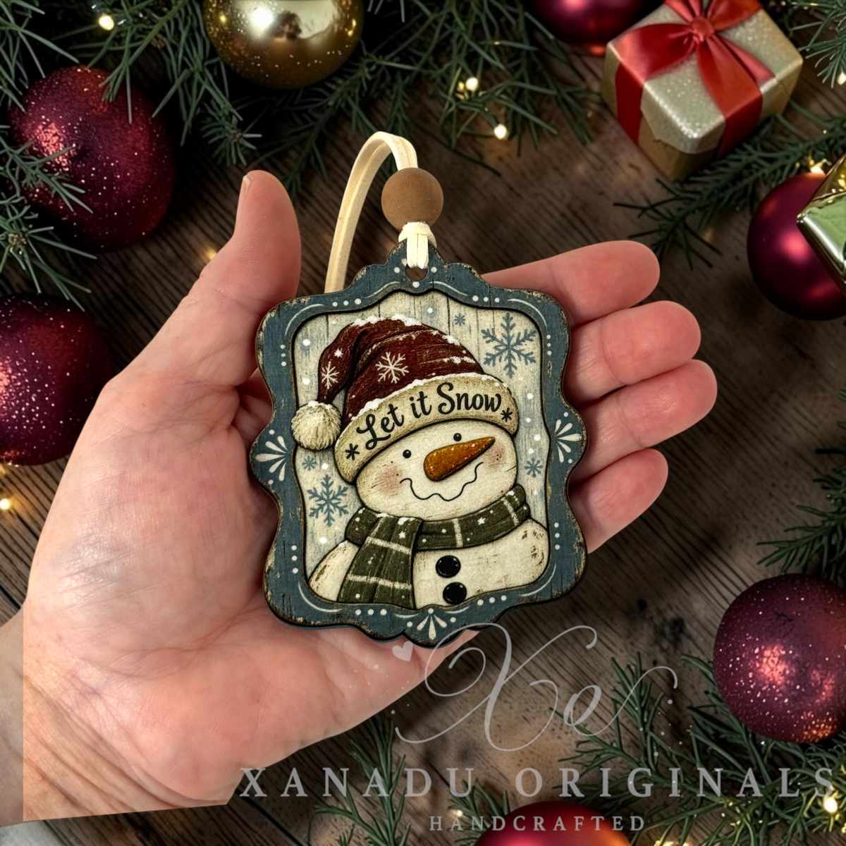 A hand holds a handcrafted wooden Christmas ornament featuring a smiling snowman wearing a maroon Santa hat and scarf with the words 'Let it Snow.' The ornament is surrounded by holiday decorations, including pine branches, red ornaments, and a gift box, on a rustic wooden surface.