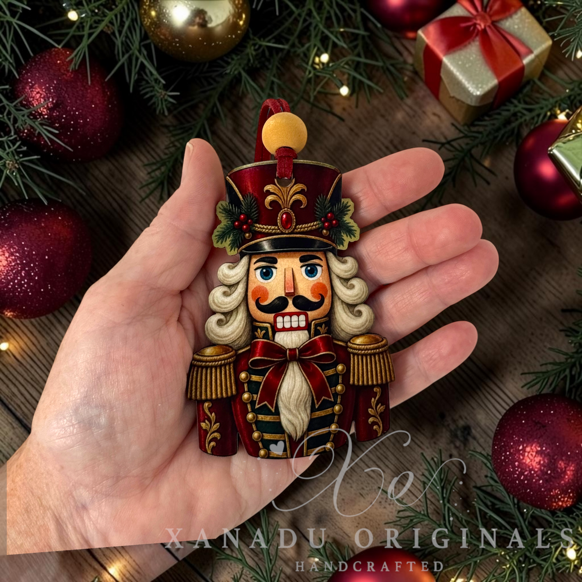 A hand holds a handcrafted wooden nutcracker ornament decorated in festive red, gold, and green colors, surrounded by Christmas decorations including pine branches, red ornaments, and a gift box.