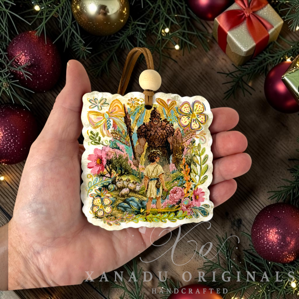 A hand holds a square, scalloped Retro religious Bible story Christmas ornament featuring an illustrated scene of a boy and a man in a forest, surrounded by colorful floral and bow motifs. The ornament is displayed against a festive holiday background with pine branches, red ornaments, and a gift box.