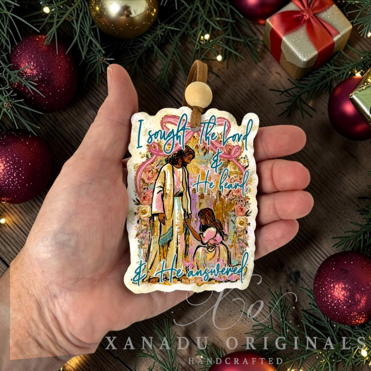 A hand holds a handcrafted Christmas ornament featuring an illustration of a woman and Jesus with the text 'I sought the Lord & He heard & He answered'. The ornament is surrounded by festive pine branches, red ornaments, and a gift box on a dark wooden surface.