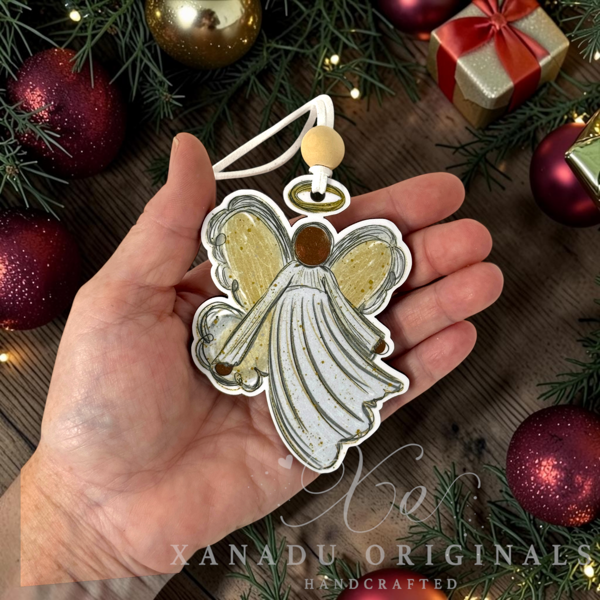 A hand holds a handcrafted angel-shaped ornament with white wings, gold glitter accents, and a white hanging loop, surrounded by Christmas decorations including red ornaments, green pine branches, and a gold gift box on a wooden surface.