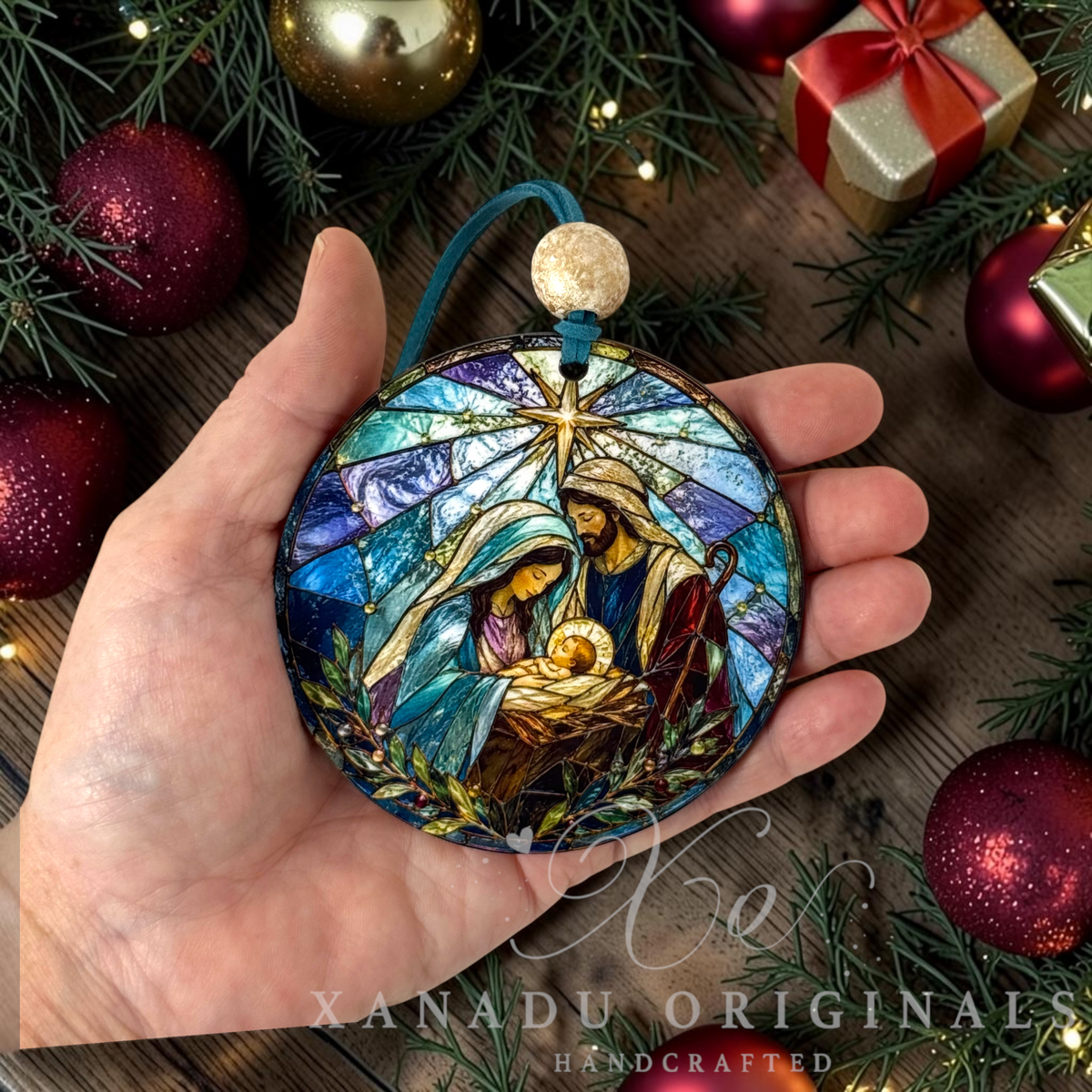 A hand holds a round stained-glass-style Christmas ornament featuring Mary, Joseph, and baby Jesus with a blue hanger and gold top. The scene is styled with pine branches, red ornaments, and a gold gift box in the background.