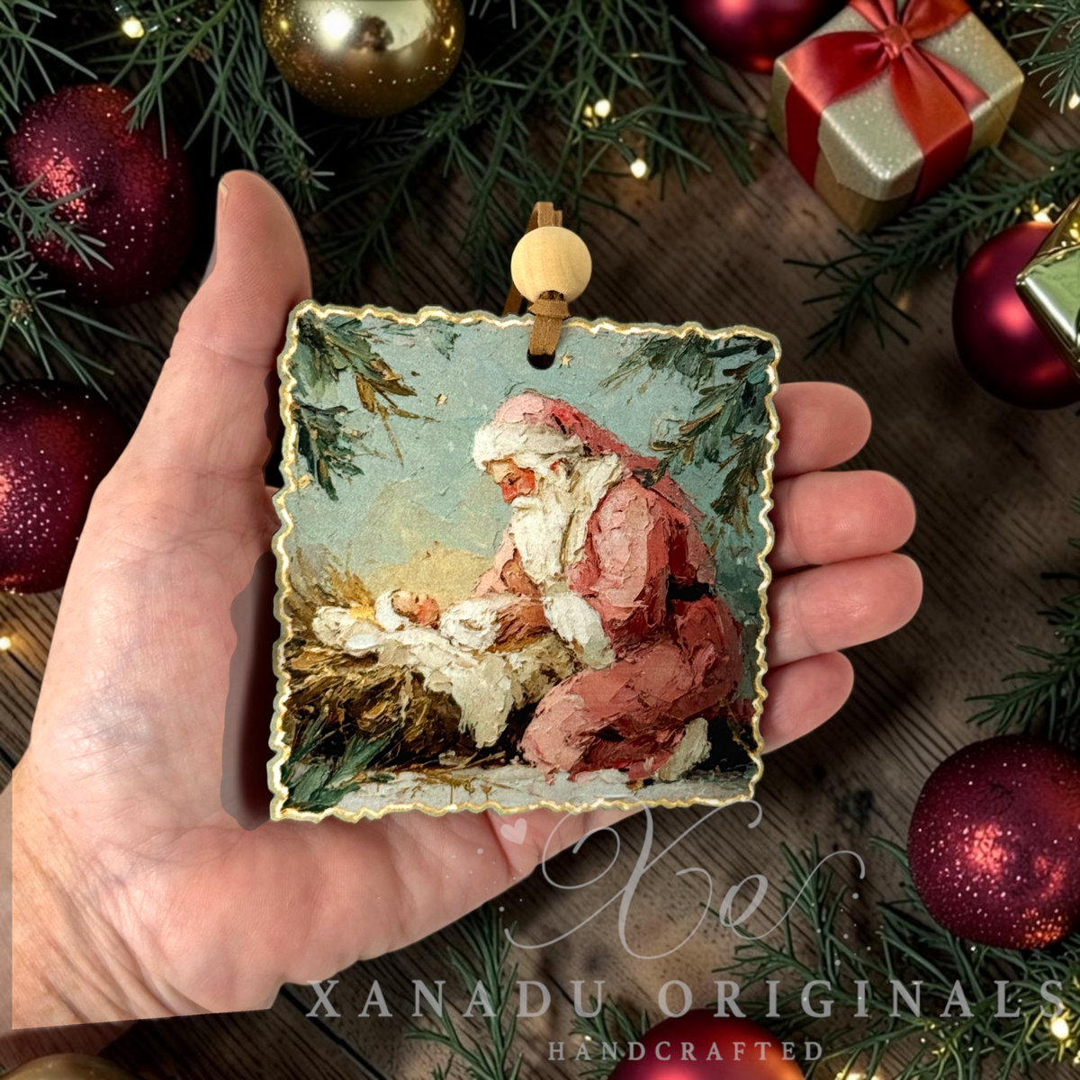 A hand holds a square scalloped-edge Christmas ornament featuring an oil painting of Santa Claus cradling baby Jesus. The ornament is decorated with a gold-toned border and a light wood hanging loop.