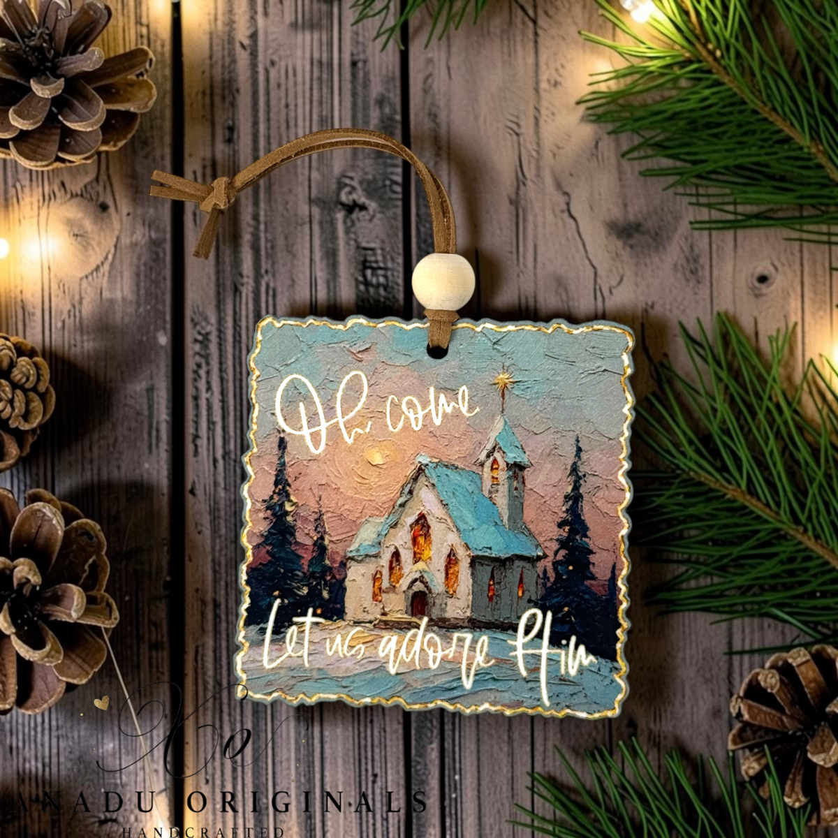 A square decorative Christmas ornament with a rustic painted church scene and the phrase āOh come Let us adore Himā in white script, hanging from a brown cord with a white bead on a wooden surface surrounded by pine branches, lights, and pinecones.