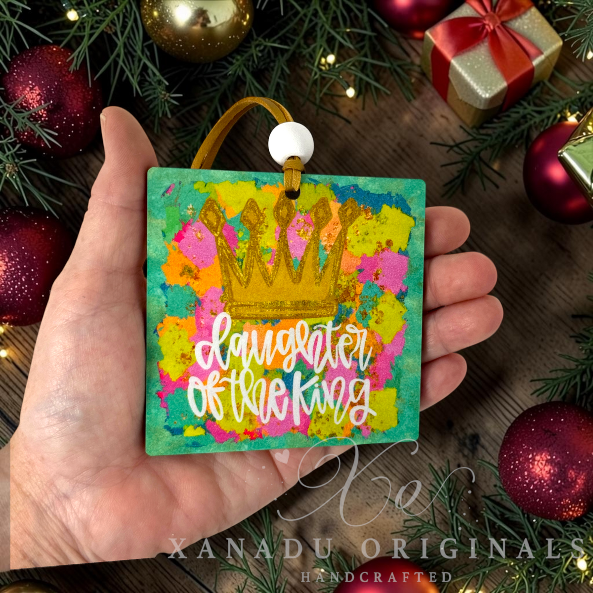 A hand holds a square wood ornament featuring a gold crown and the text 'daughter of the king' in white script over a colorful splattered background. The scene is decorated with pine branches, red ornaments, and a gold gift box, with a 'XANADU ORIGINALS HANDCRAFTED' watermark at the bottom.