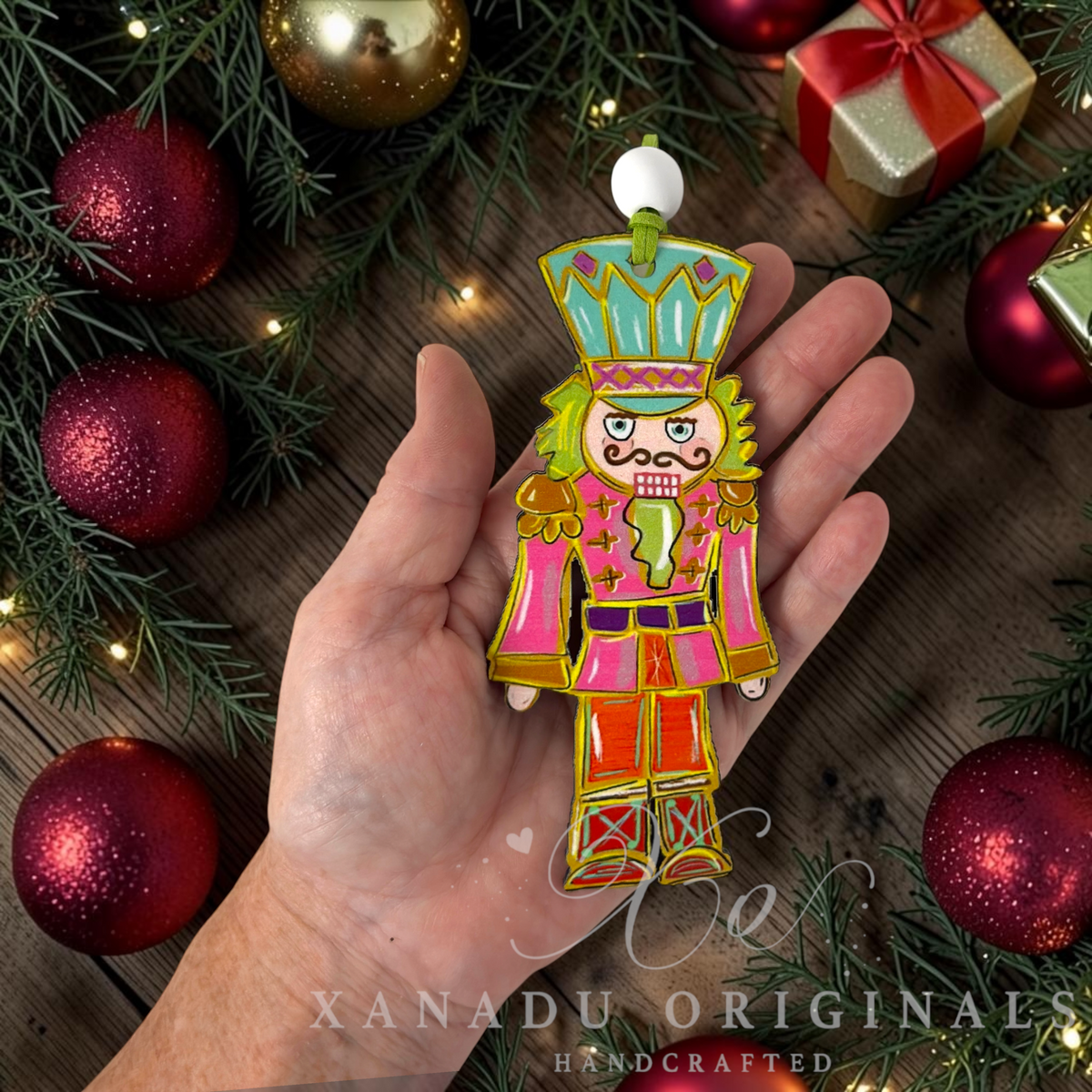 A hand holds a colorful hand-painted nutcracker ornament against a rustic wooden background decorated with pine branches, red ornaments, and a gold gift box. The ornament features a pink jacket, red pants, and a green and blue hat.