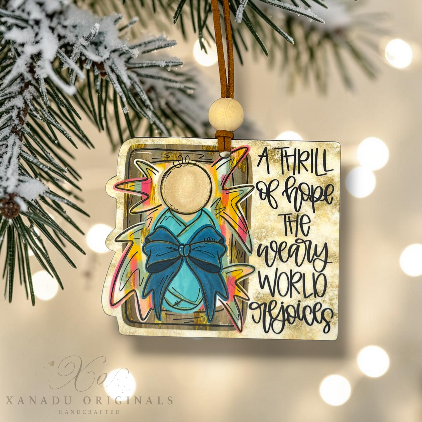 A square acrylic Christmas ornament hanging from a brown string with a wooden bead. The ornament features a stylized angel figure in blue with a pink and yellow halo, alongside handwritten text reading 'A THRILL of hope the every WORLD around us.