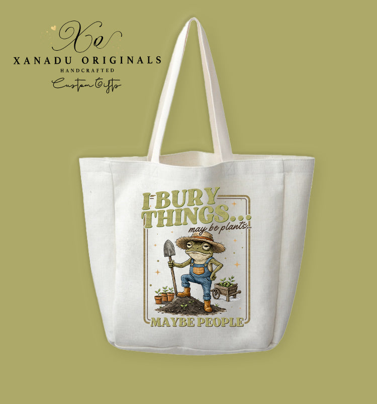 A cream-colored polyester linen tote bag featuring a retro framed watercolor illustration of a frog in overalls and a straw hat holding a garden trowel on a mound of dirt. Distressed green and gold text reads "I BURY THINGS... may be plants... MAYBE PEOPLE."