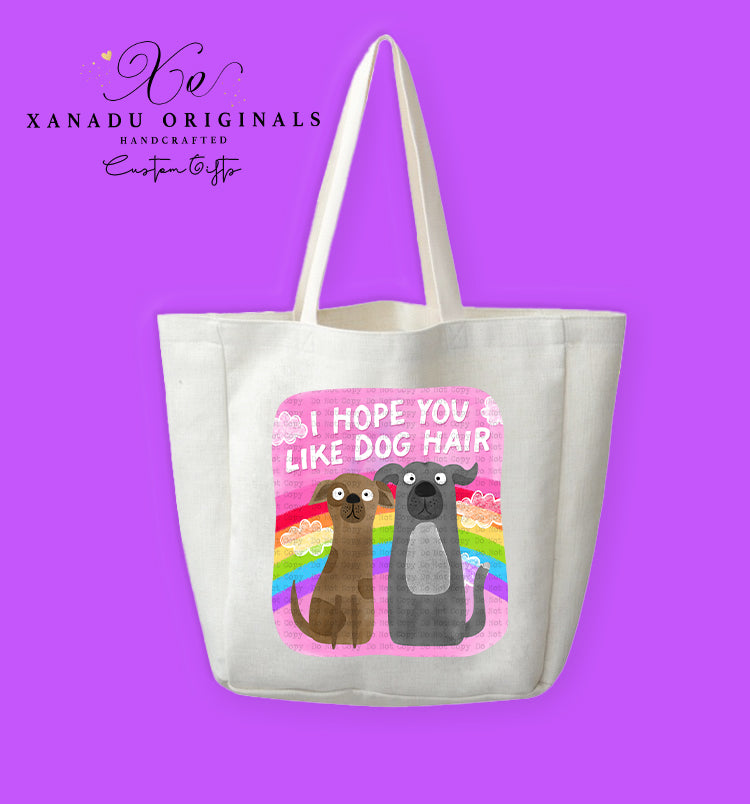 A cream-colored polyester linen tote bag featuring a square pink graphic with a retro rainbow and white clouds. In front sit two wide-eyed cartoon dogs (one brown, one grey) below bold white text that reads "I HOPE YOU LIKE DOG HAIR."