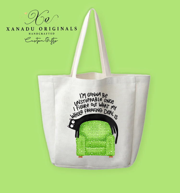 A cream-colored polyester linen tote bag featuring a cartoon illustration of a wide-eyed black cat draped over the back of a bright green armchair with small white patterns. Bold black text arched above the graphic reads "I'M GONNA Be UNSTOPPABLE ONCE I FiGURE OUT WHAT MY WHOLE FREAKING DEAL IS."
