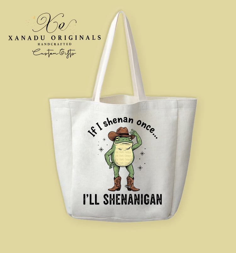 A cream-colored polyester linen tote bag featuring a whimsical cartoon illustration of a green frog wearing a brown cowboy hat and cowboy boots. The frog is tipping his hat amidst small black sparkle stars, framed by text reading "If I shenan once... I'LL SHENANIGAN."