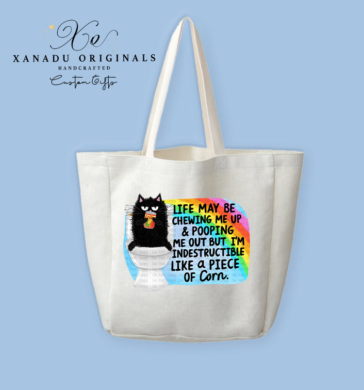 A cream-colored polyester linen tote bag featuring a cartoon illustration of a grumpy black cat sitting on a toilet eating from a can labeled "CORN." The background is a bright rainbow cloud, with bold black text that reads "LIFE MAY BE CHEWING ME UP & POOPING ME OUT BUT I'M INDESTRUCTIBLE LIKE A PIECE OF CORN."