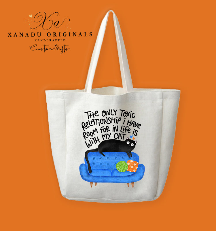 A cream-colored polyester linen tote bag featuring a cartoon illustration of a wide-eyed black cat lounging on top of a bright blue sofa with decorative green and orange pillows. Bold black text above the graphic reads "THe oNLY Toxic ReLaTIoNSHIP i HaVe RooM foR iN LiFe iS WITH MY CAT."