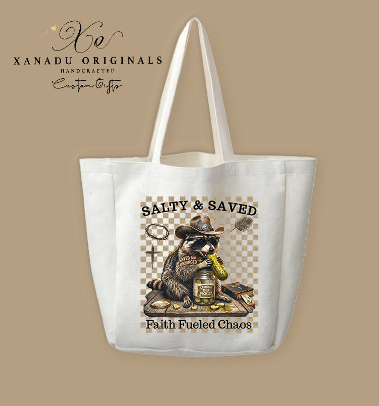A cream-colored polyester linen tote bag featuring a rustic western illustration of a raccoon in a cowboy hat eating pickles with the text "SALTY & SAVED" and "Faith Fueled Chaos".