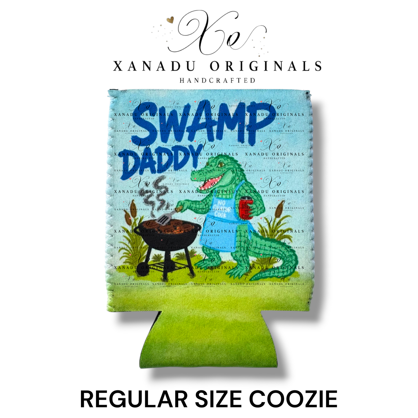 "Xanadu Originals handcrafted regular size neoprene can coozie featuring an illustration of a green alligator wearing a blue apron while grilling on a barbecue with the bold blue text 'SWAMP DADDY' over a scenic bayou background."