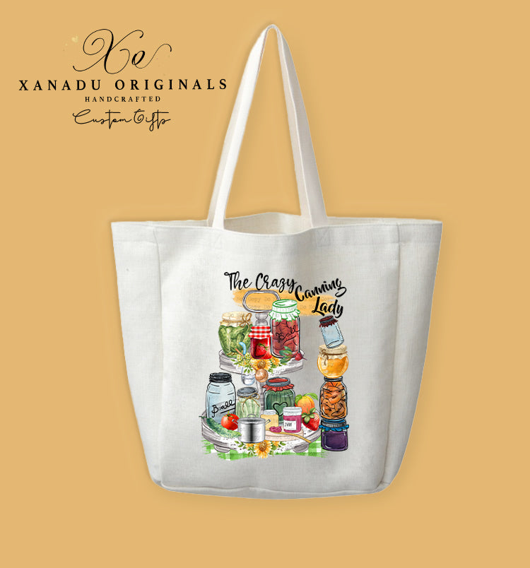 A cream-colored polyester linen tote bag featuring a watercolor illustration of a multi-tiered display stand holding numerous colorful jars of canned preserves and pickles, accented with sunflowers and green plaid fabric under black text reading "The Crazy Canning Lady."