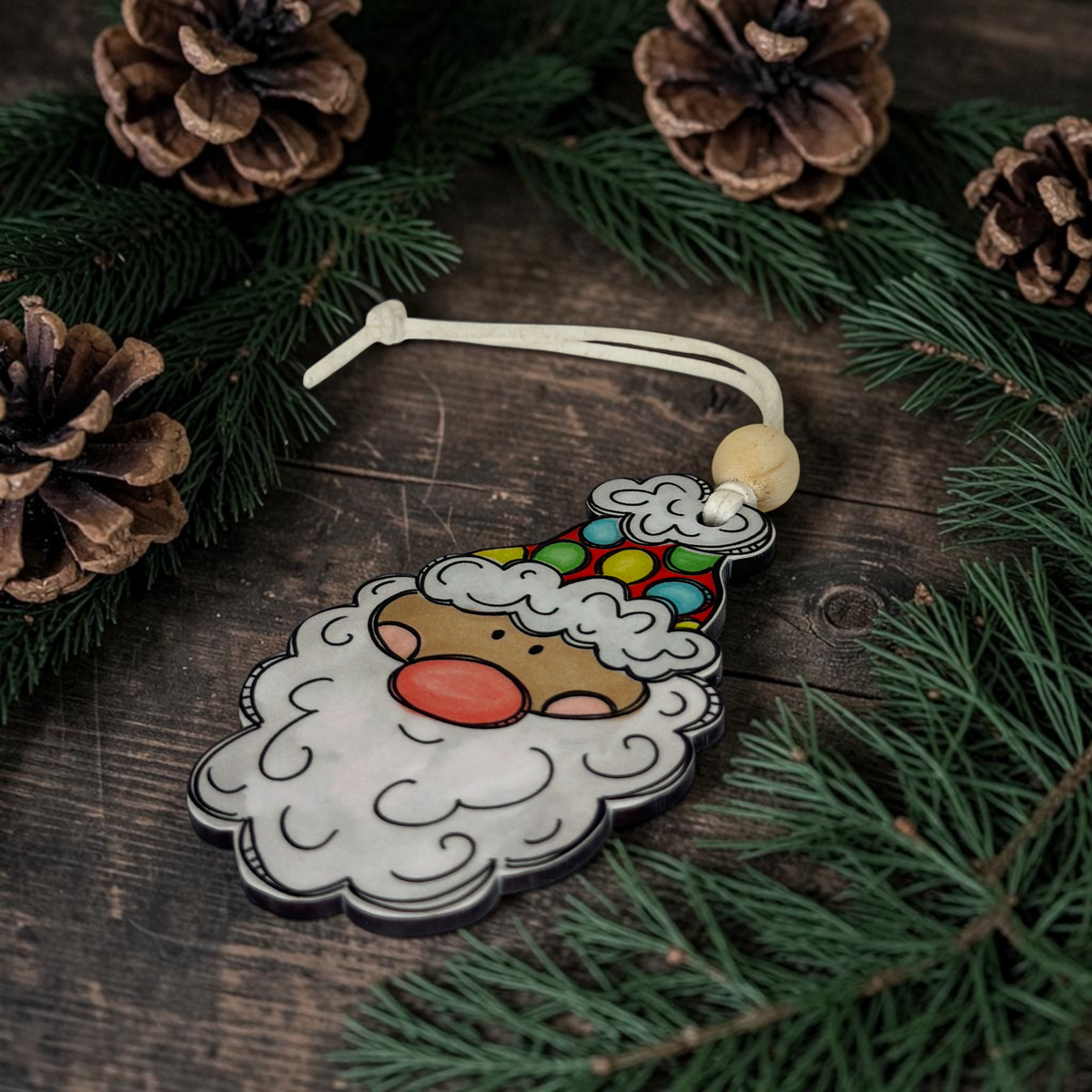 A light skin tone retro Santa face wood ornament with a bright polka dot hat and a natural wood bead, resting on a dark plank wood background with rustic pine cones.