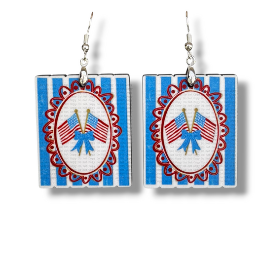 A pair of square MDF earrings featuring a vintage-style illustration of two crossed American flags tied with a blue bow. The flags are centered in a red scalloped oval frame over a blue and white vertical striped background, hanging from silver hooks.